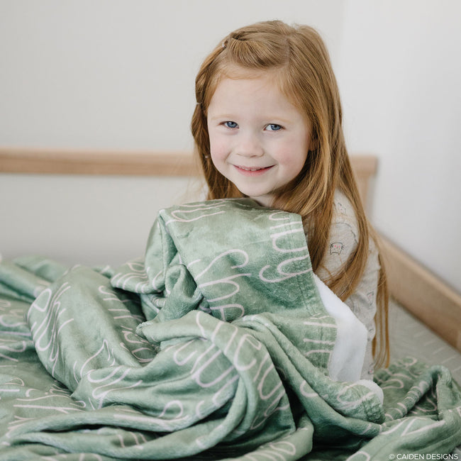 A young girl with a green personalized blanket with the name 'Shelby' printed in a blush font.