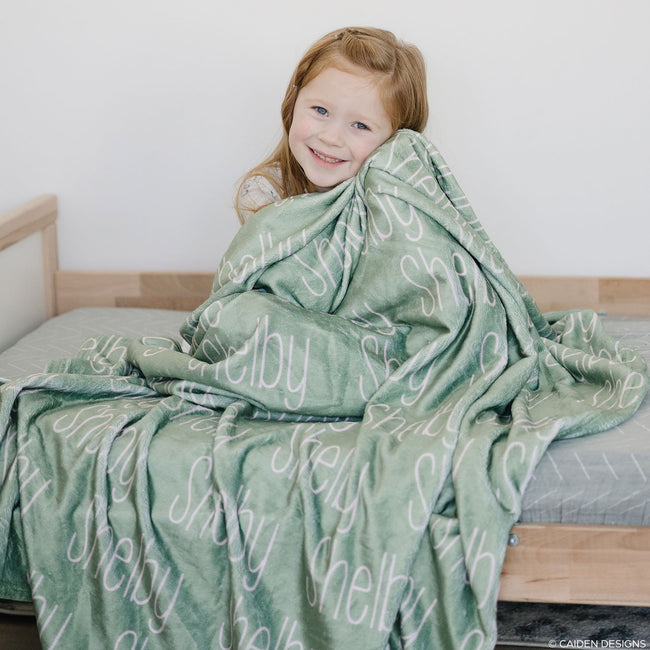 A young girl with a sage personalized blanket with the name 'Shelby' printed in a blush font.