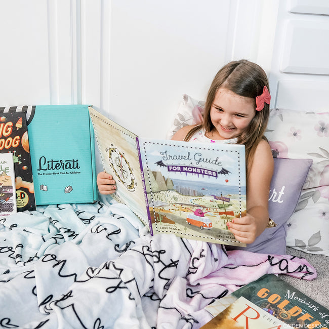 A child sitting on a bed reading a book, covered with a personalized blanket that has a pastel ombre design.