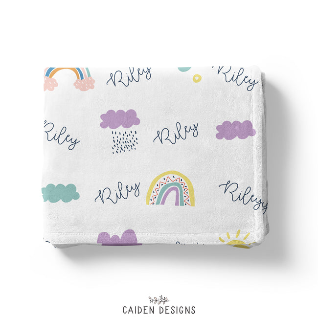 A personalized blanket with a rainbows, clouds, and suns pattern and the name 'Riley' printed repeatedly.