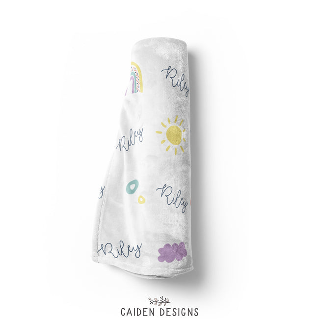 A rolled personalized blanket with a rainbows, clouds, and suns pattern and the name 'Riley' printed repeatedly.