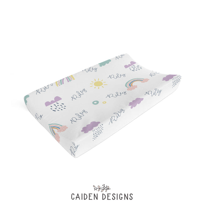 A personalized changing pad cover with a Scandinavian rainbow and cloud pattern, lying flat on a surface.