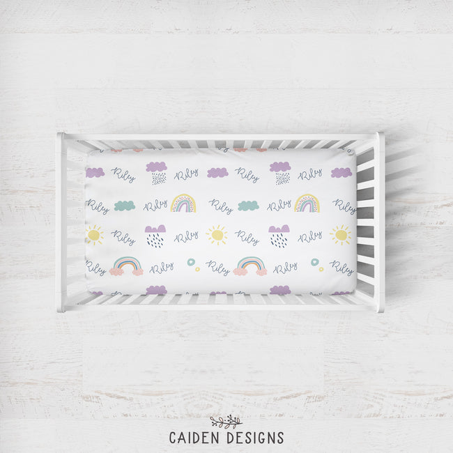 A personalized crib sheet with Scandinavian rainbows design, featuring names in purple and yellow text, displayed in a white crib.