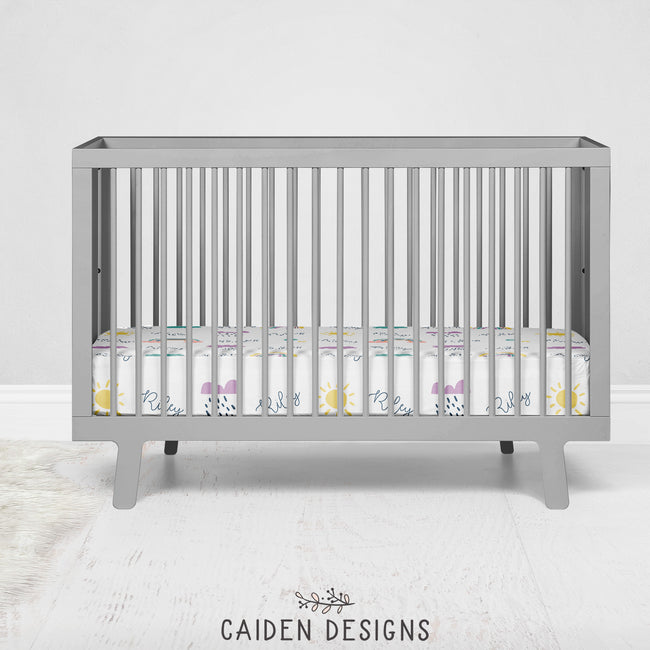 A personalized crib sheet with Scandinavian rainbows design, featuring names in purple and yellow text, displayed in a grey crib.