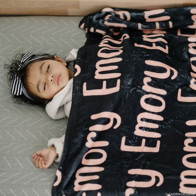 A baby sleeping peacefully under a personalized blanket with the name 'Emory' printed in a repeating pattern across the fabric.