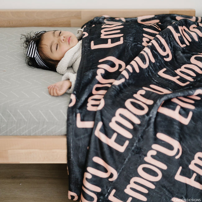 A baby sleeping peacefully under a personalized blanket with the name 'Emory' printed in a repeating pattern across the fabric.