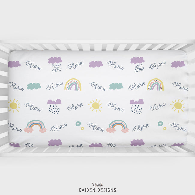 A personalized crib sheet with Scandinavian rainbows design, featuring names in purple and yellow text, displayed in a white crib.