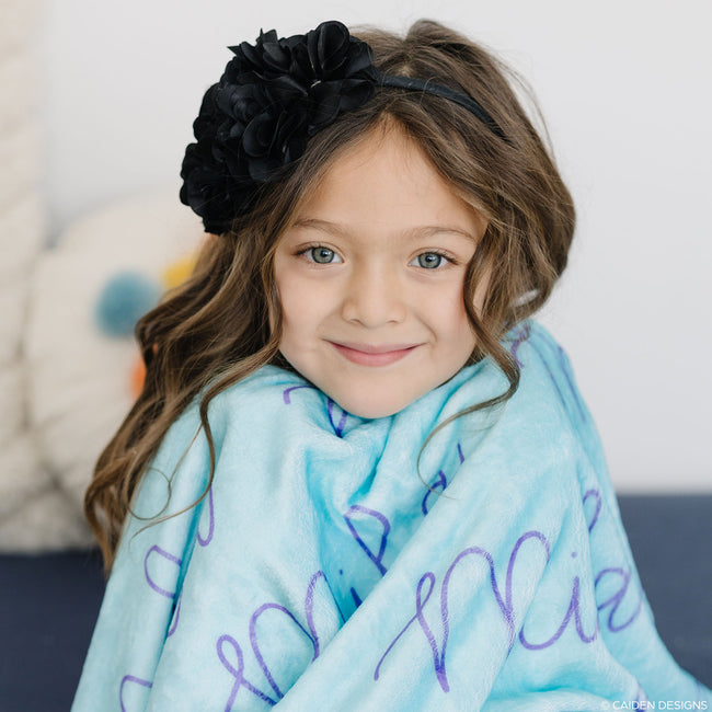 A child with an aqua blanket with the name 'Mila' printed in royal purple text.