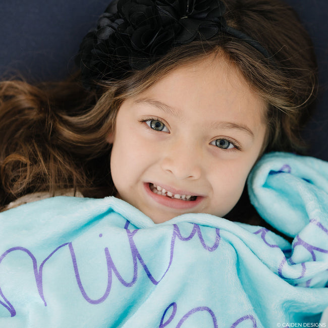 A child with a smile on their face lying on a aqua blanket with the name 'Mila' printed in royal purple text.