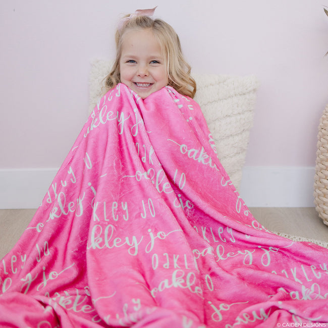 A smiling child wrapped in a pink personalized blanket with soft mint text.