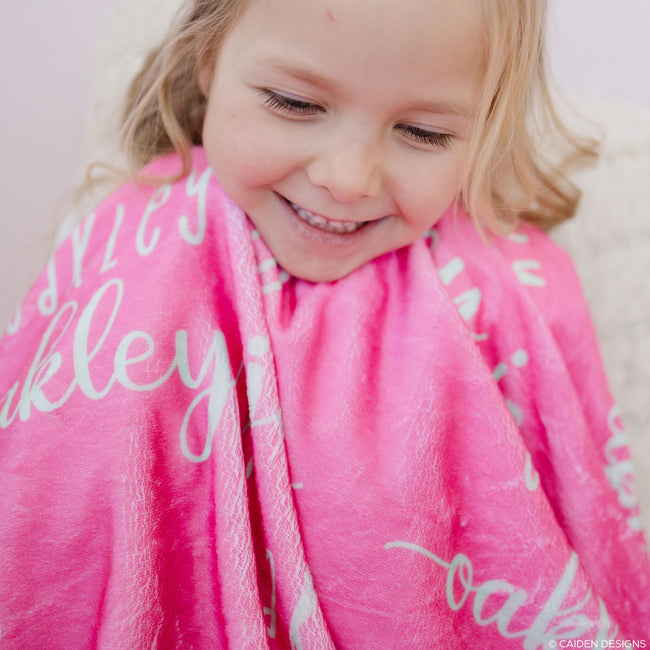 A smiling child wrapped in a pink personalized blanket with soft mint text.