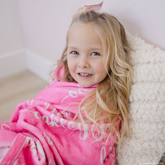 A smiling child wrapped in a pink personalized blanket with soft mint text.