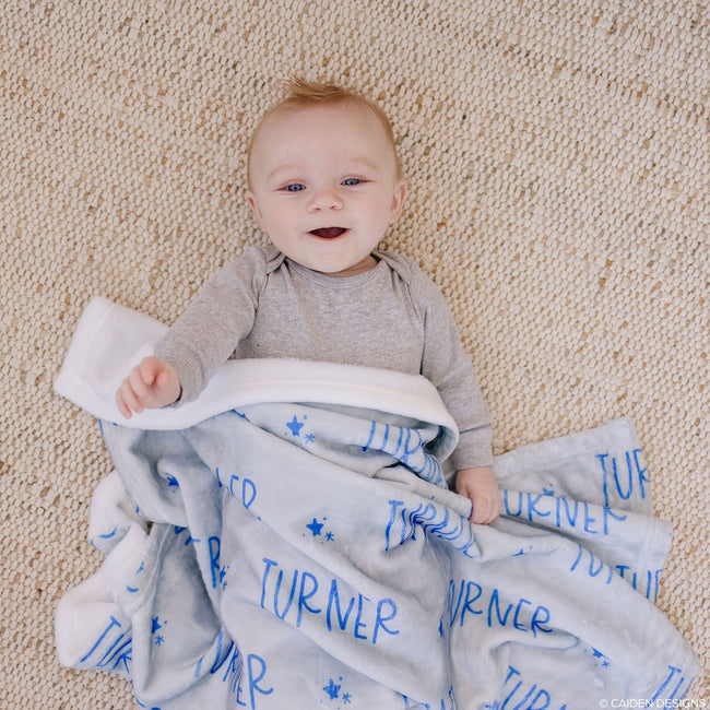 A baby lying on a dusty blue personalized blanket with the name 'Turner' printed on it.