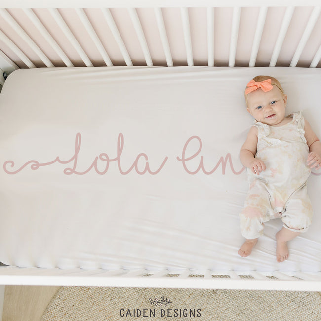 A personalized crib sheet with the name 'Lola Ann' printed in a cursive script on a crib, with a baby lying on the crib