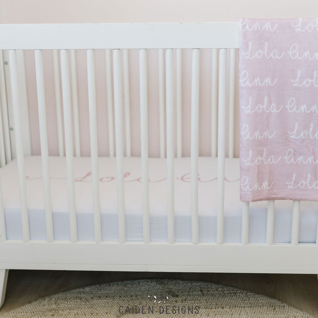 A personalized crib sheet with the name 'Lola Ann' printed in a cursive script on a crib and a matching personalized baby blanket