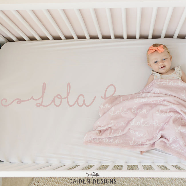 A personalized crib sheet with the name 'Lola' printed in a cursive script on a crib, with a baby lying on the crib