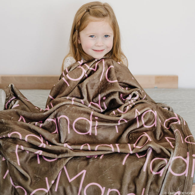 Little girl sitting on a bed with a brown personalized blanket with names on it