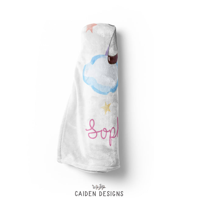 A rolled personalized unicorn blanket with the name 'Sophia Grace' printed on it, featuring a colorful unicorn design and other playful elements like stars and a rainbow.