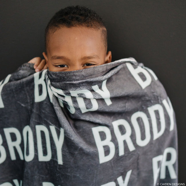 a little boy hiding behind a charcoal personalized blanket with the name Brody