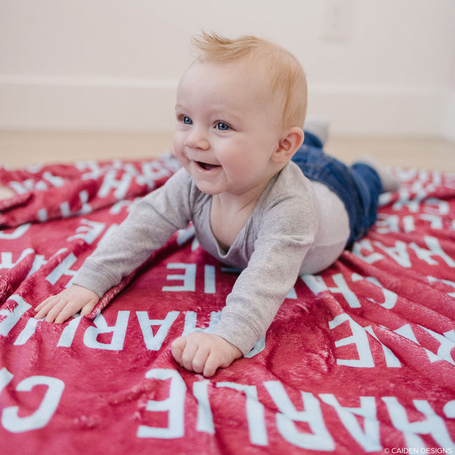 A baby lying on a red personalized blanket with the name 'Charlie' printed in white text