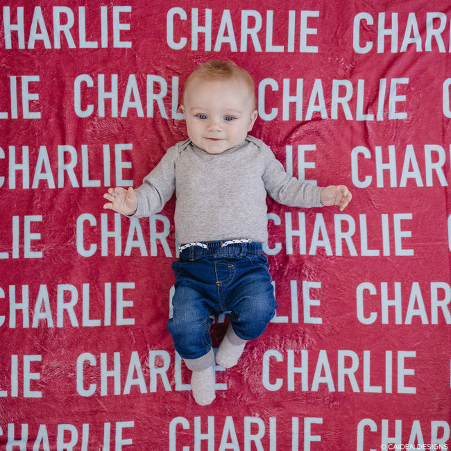 A baby lying on a red personalized blanket with the name 'Charlie' printed in white text
