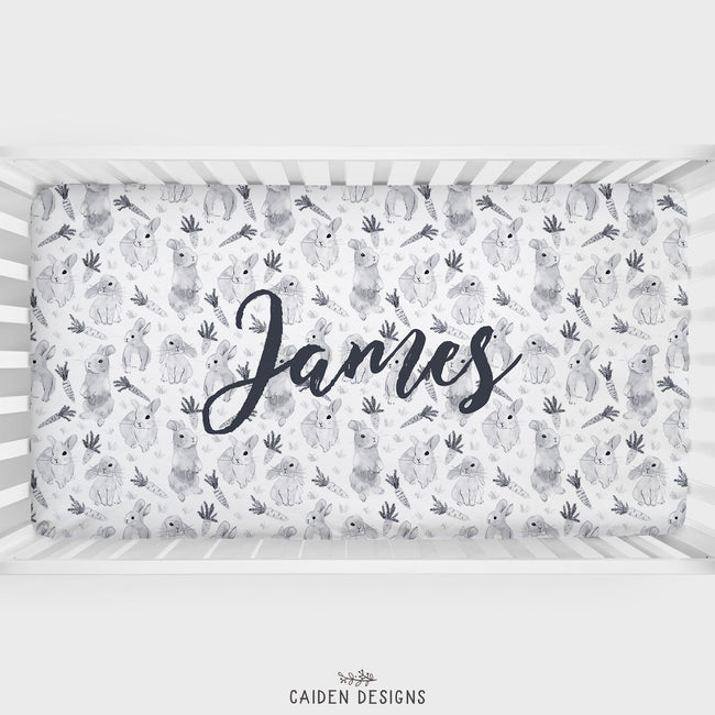 A gray and white watercolor bunny crib sheet with the name 'James' personalized on it, displayed in a crib.