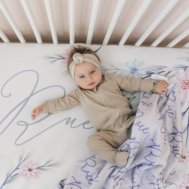 A baby lying on a crib with a personalized crib sheet featuring pastel watercolor flowers