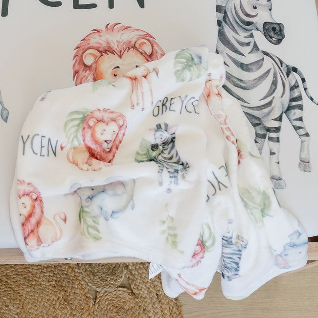 A personalized white blanket featuring repeated watercolor giraffe, elephant, lion, zebra and plants with the name "GREYCEN" in black, capitalized letters. 