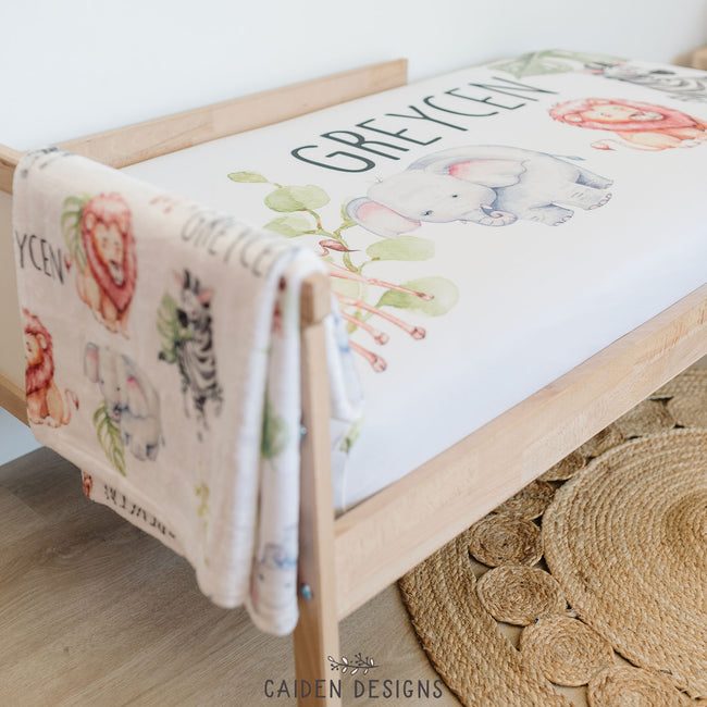 A personalized white blanket featuring repeated watercolor giraffe, elephant, lion, zebra and plants with the name "GREYCEN" in black, capitalized letters. A matching crib sheet sits in the background.