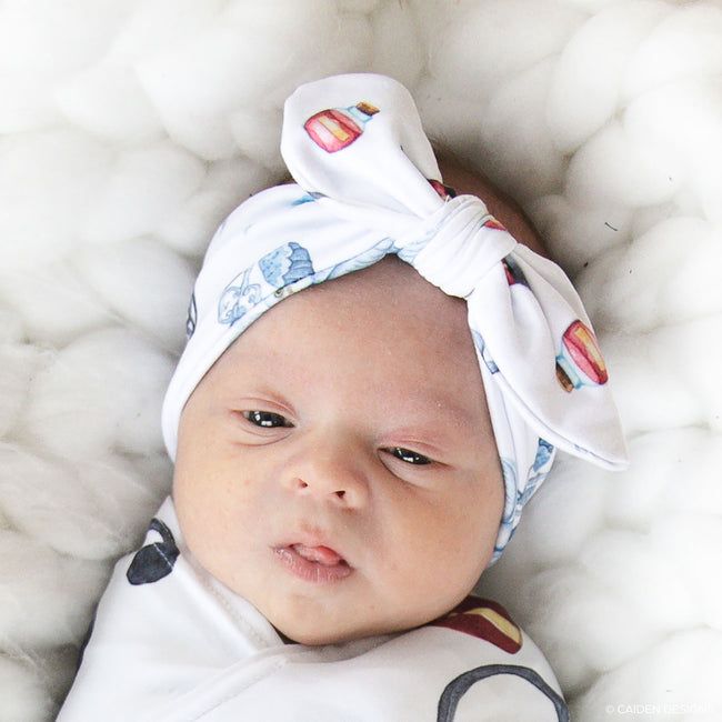 A swaddled baby with a wizard design, featuring a blue and white knit pattern with personalized text, lying in a wicker bassinet.