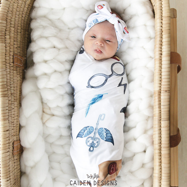 A swaddled baby with a wizard design, featuring a blue and white knit pattern with personalized text, lying in a wicker bassinet.