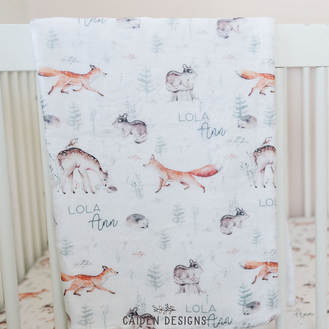 a personalized baby blanket featuring a woodland animals design draped over a crib 